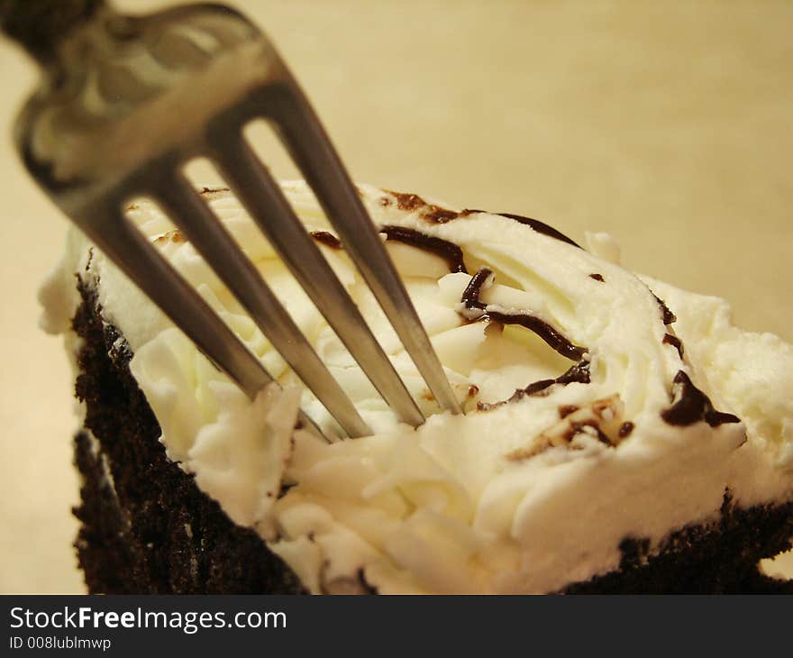 A large wedge of chocolate cake with white icing and white chocolate shavings on top with a fork digging in for a bite. A large wedge of chocolate cake with white icing and white chocolate shavings on top with a fork digging in for a bite.