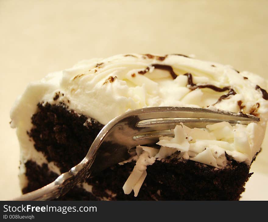 A large wedge of chocolate cake with white icing and white chocolate shavings on top with a fork digging in for a bite. A large wedge of chocolate cake with white icing and white chocolate shavings on top with a fork digging in for a bite.