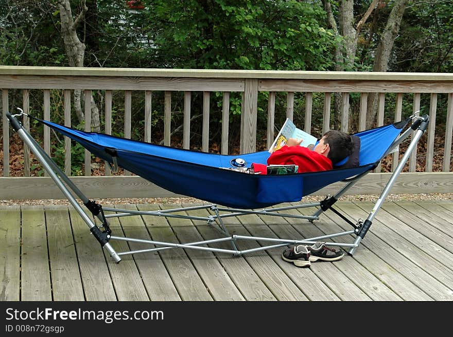 A young boy is laying in a blue hammock reading a book. A young boy is laying in a blue hammock reading a book.