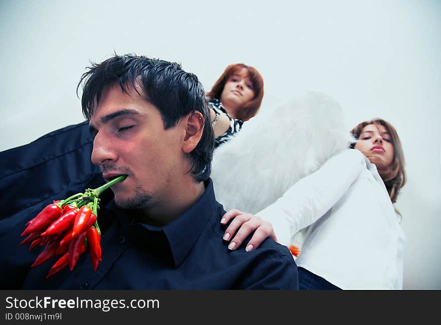 Two beautiful young girls with angel wings holding hands on a boy sitting in his knees and holding a buch of red chilli peppers in his mouth. Two beautiful young girls with angel wings holding hands on a boy sitting in his knees and holding a buch of red chilli peppers in his mouth