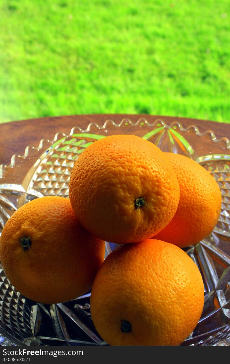 Four juicy, ripe oranges in a glass, crystal bowl on a round, polished, wooden table with lush green grass and pale yellow curtains in the background. Four juicy, ripe oranges in a glass, crystal bowl on a round, polished, wooden table with lush green grass and pale yellow curtains in the background