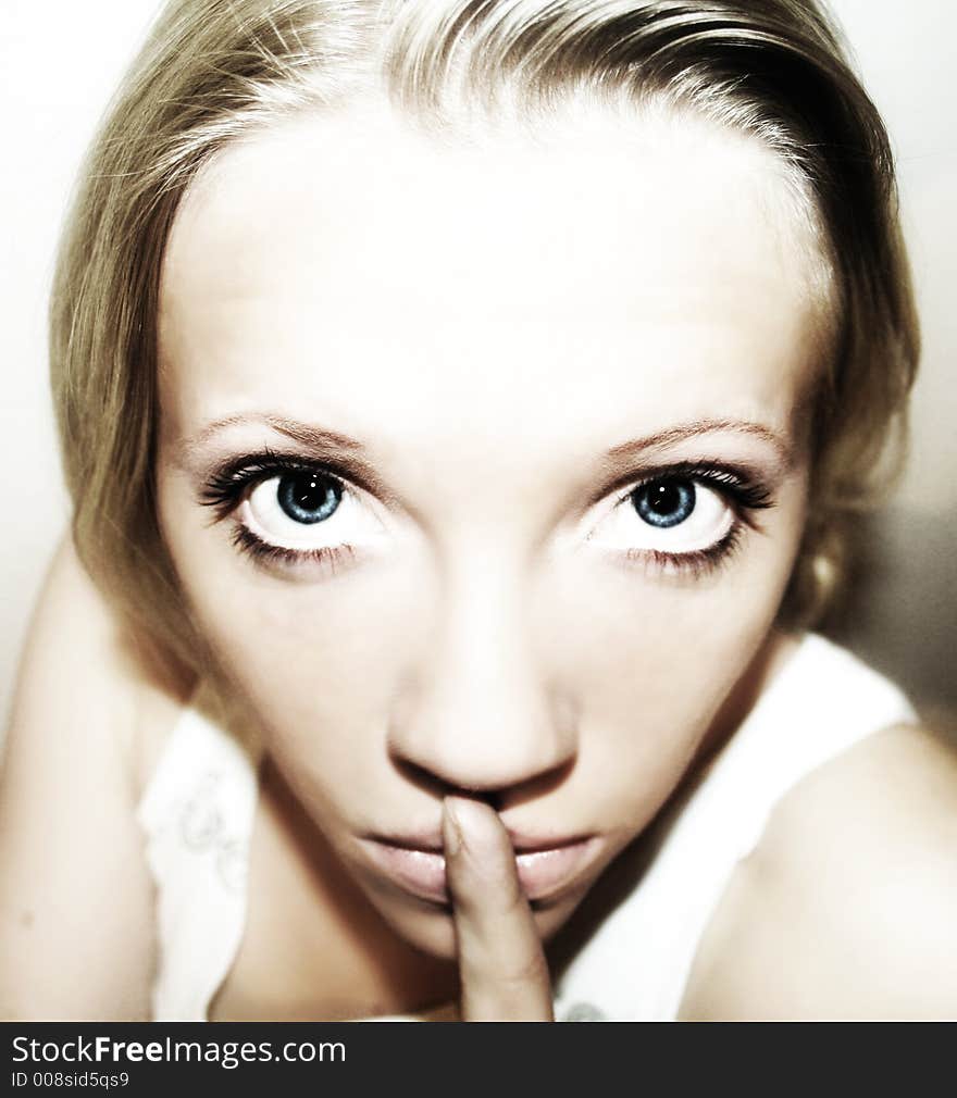 A young blond woman with blue eyes in front of white background is holding her finger in front of her mouth. A young blond woman with blue eyes in front of white background is holding her finger in front of her mouth