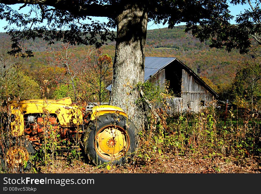 Old tractor sets beside tree with a barn in the distance.  The tractor is overgrown with weeds and is yellow. Old tractor sets beside tree with a barn in the distance.  The tractor is overgrown with weeds and is yellow.