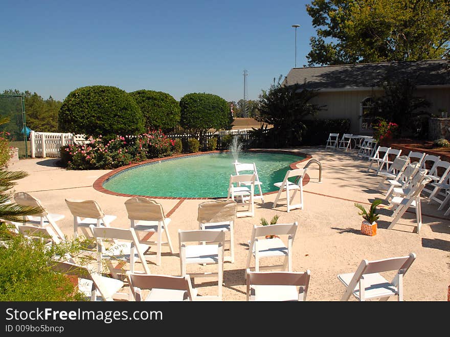 A beautiful blue green pool with a flowing fountain. It's surrounded with white chairs waiting for the wedding guests. A beautiful blue sky, white pickett fence and beautiful blooming flowers make the setting a perfect site for the wedding. A beautiful blue green pool with a flowing fountain. It's surrounded with white chairs waiting for the wedding guests. A beautiful blue sky, white pickett fence and beautiful blooming flowers make the setting a perfect site for the wedding.