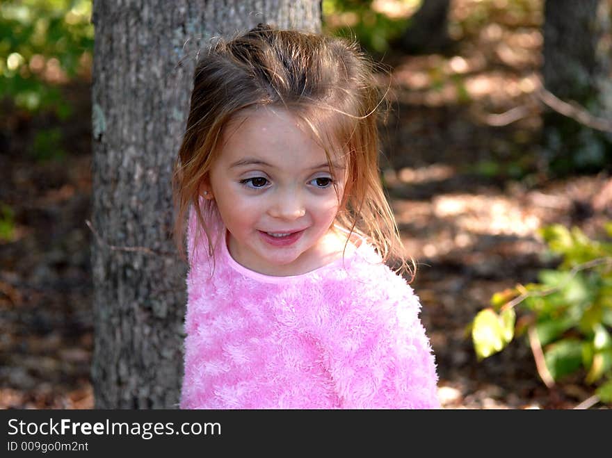 Beautiful little girl standing outdoors by a tree. She's smiling and looking as something has her attention. She's wearing a pink fuzzy shirt. Beautiful little girl standing outdoors by a tree. She's smiling and looking as something has her attention. She's wearing a pink fuzzy shirt.