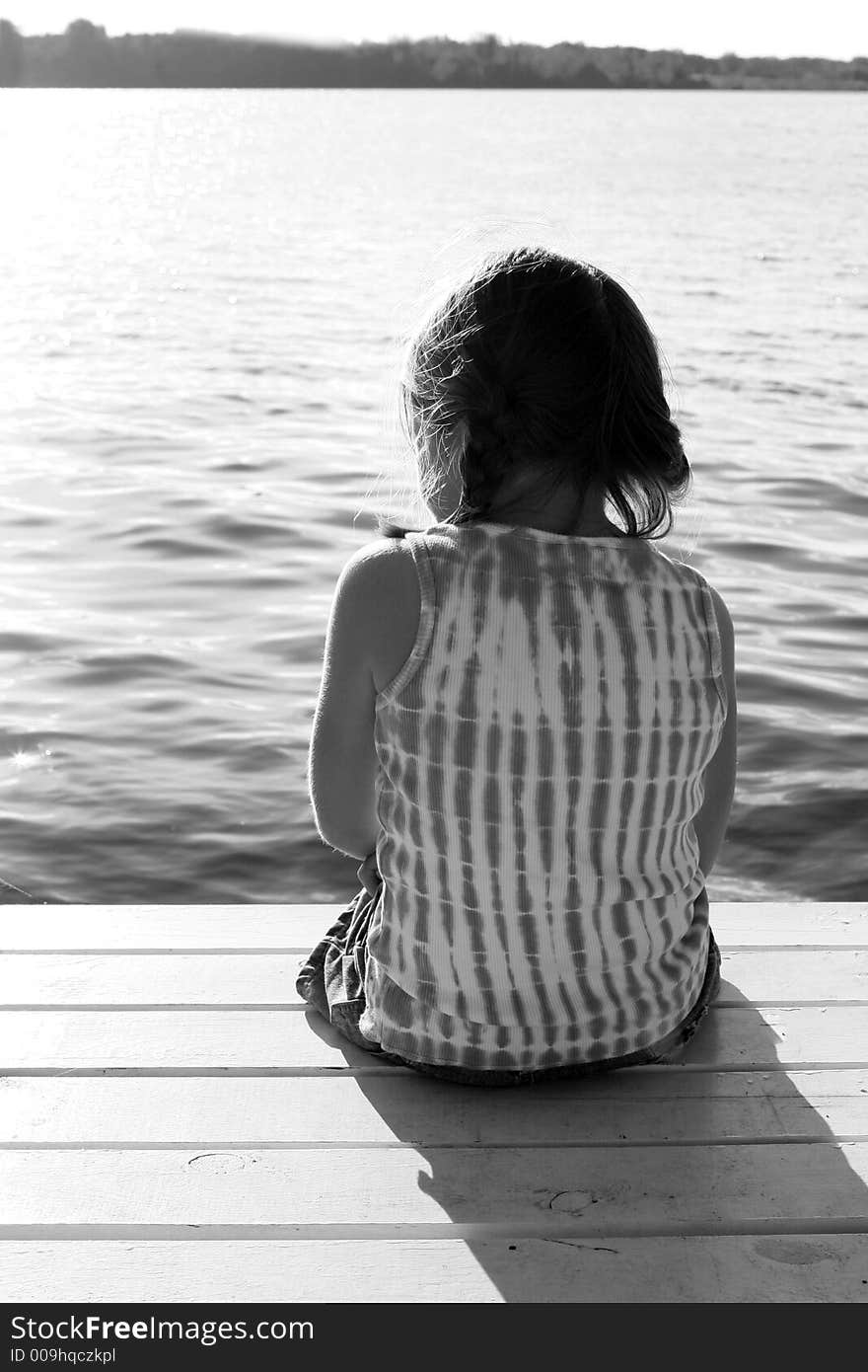 Black and white image of a girl sitting on a pier, overlooking a lake. Black and white image of a girl sitting on a pier, overlooking a lake