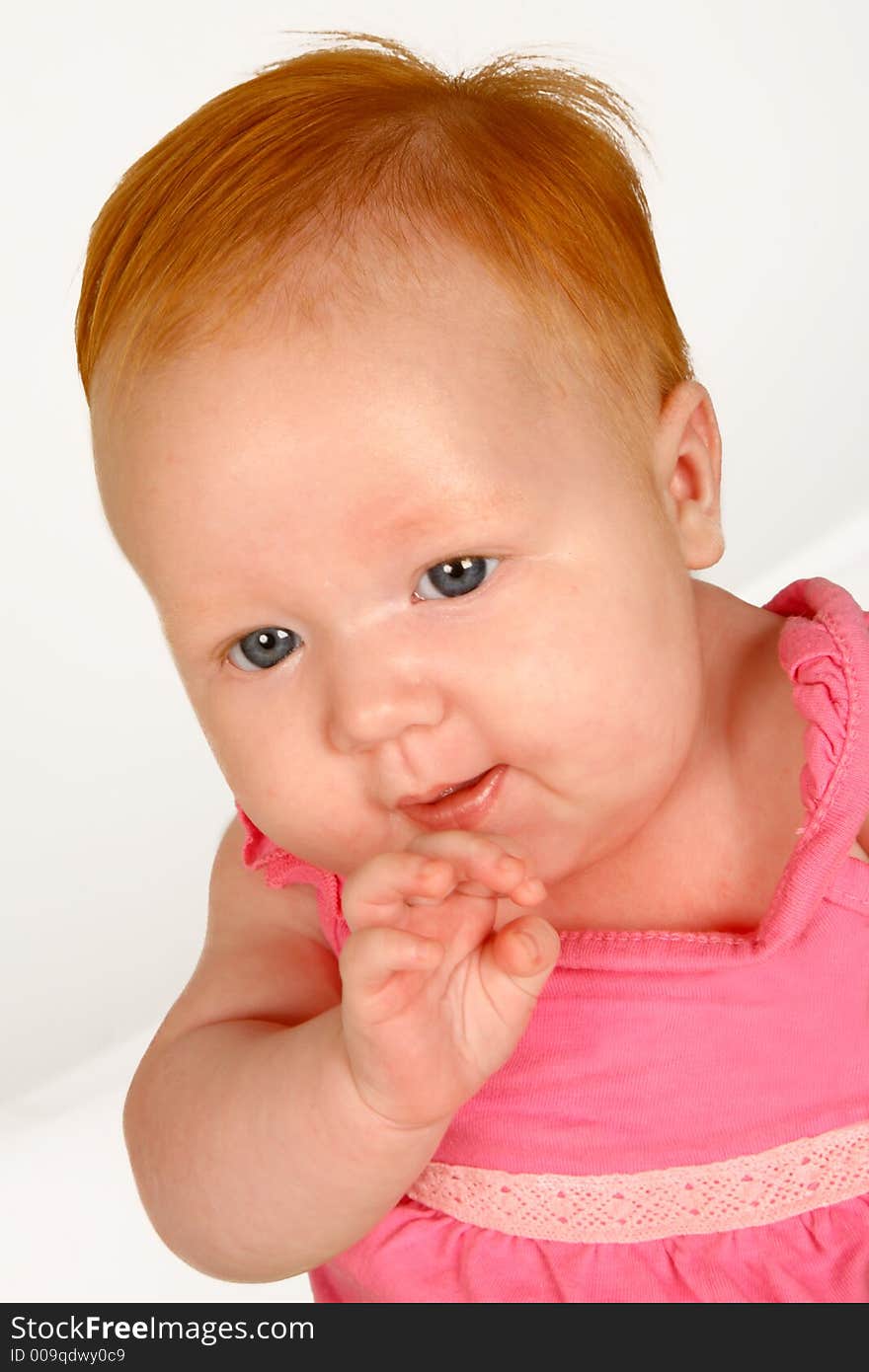 A baby girl sitting holding her hand by her mouth. A baby girl sitting holding her hand by her mouth