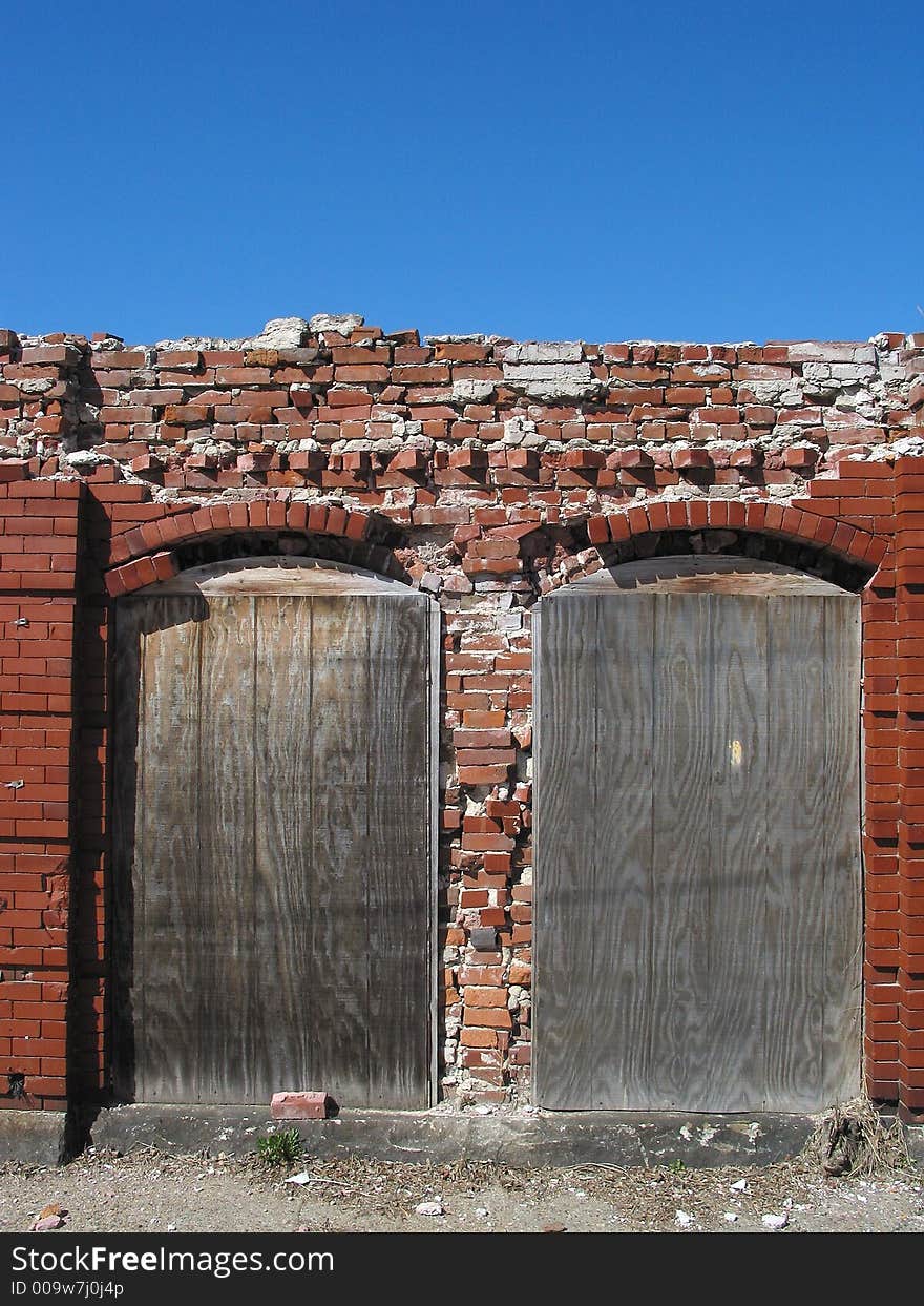 Description: Industrial ruins of two boarded-up doors framed by bricks.  There is a blue sky in the top third of the photo.  The top portion of the bricks are crumbling. Description: Industrial ruins of two boarded-up doors framed by bricks.  There is a blue sky in the top third of the photo.  The top portion of the bricks are crumbling.