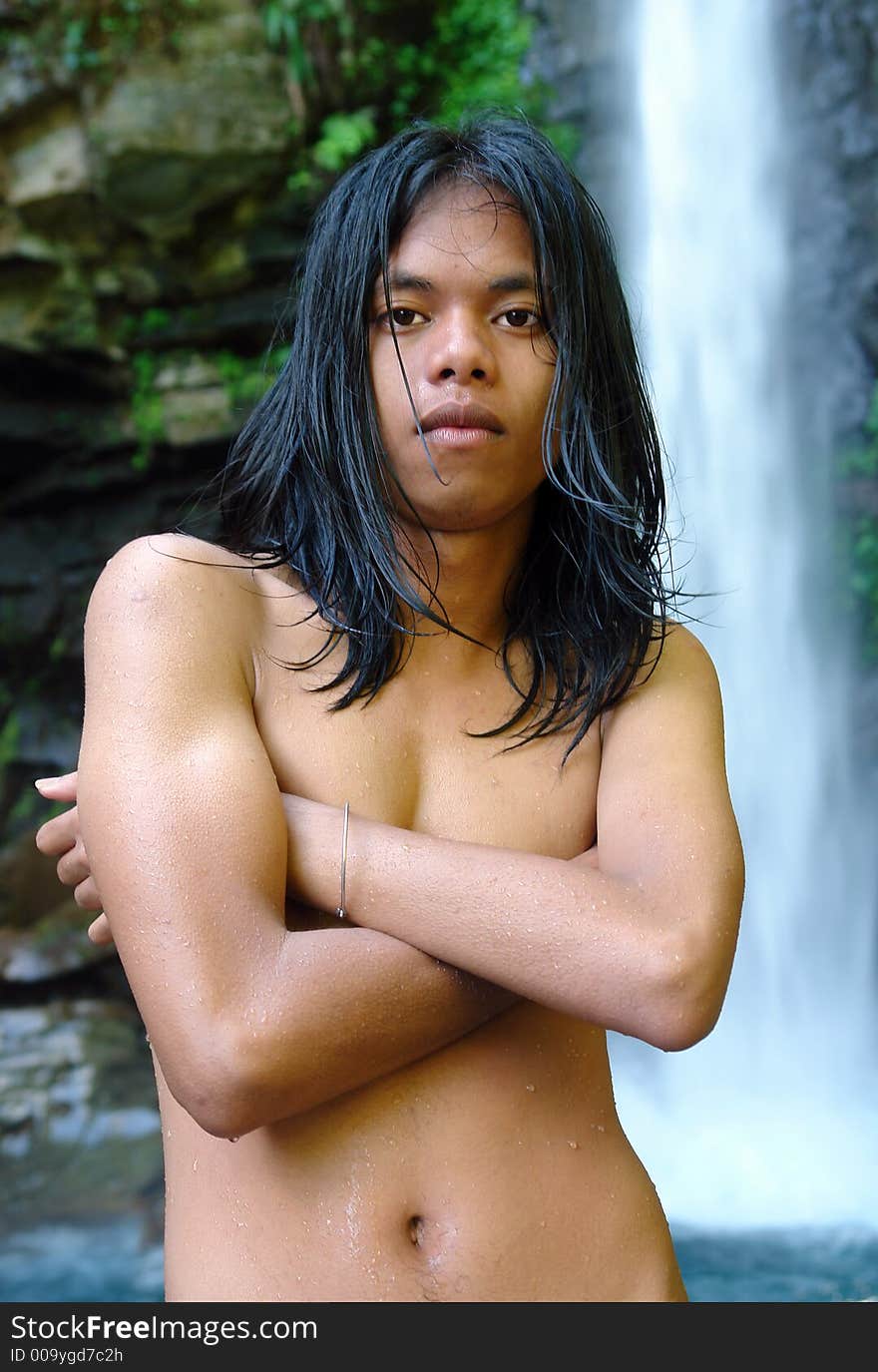 Asian exotic long-haired boy in front of a tropical pristine waterfall, after taking a dip in the whirlpool, shivering and covered with droplets. Asian exotic long-haired boy in front of a tropical pristine waterfall, after taking a dip in the whirlpool, shivering and covered with droplets.