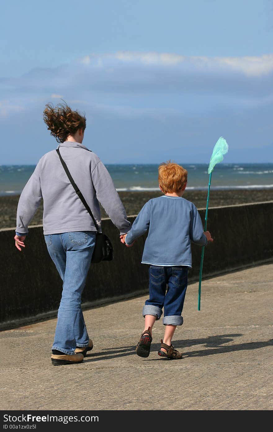 Woman holding the hand of a young boy holding a green fishing net in his hand and walking along a beach promenade. Sea out of focus to the rear. Woman holding the hand of a young boy holding a green fishing net in his hand and walking along a beach promenade. Sea out of focus to the rear.