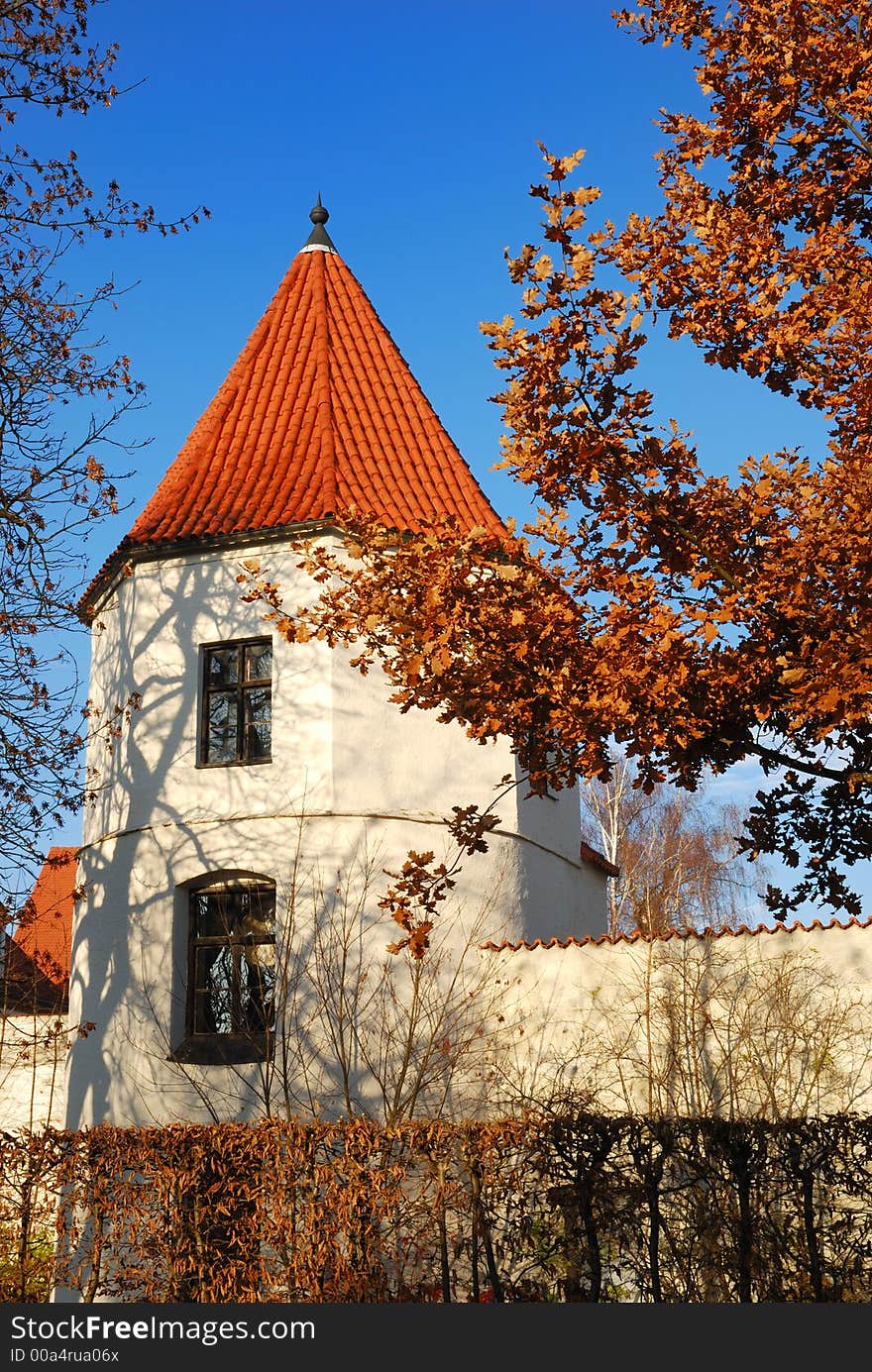 The image shows an old, historic tower of a city wall, seen in a small Bavarian town. The tower has a white wall with two windows and a red rood. The shadow of a tree can be seen on the building. The picture is framed by two trees with already brown leaves. The shot was taken on a sunny autumn day. The sky is deep blue. In front of the fortification is a hedge.