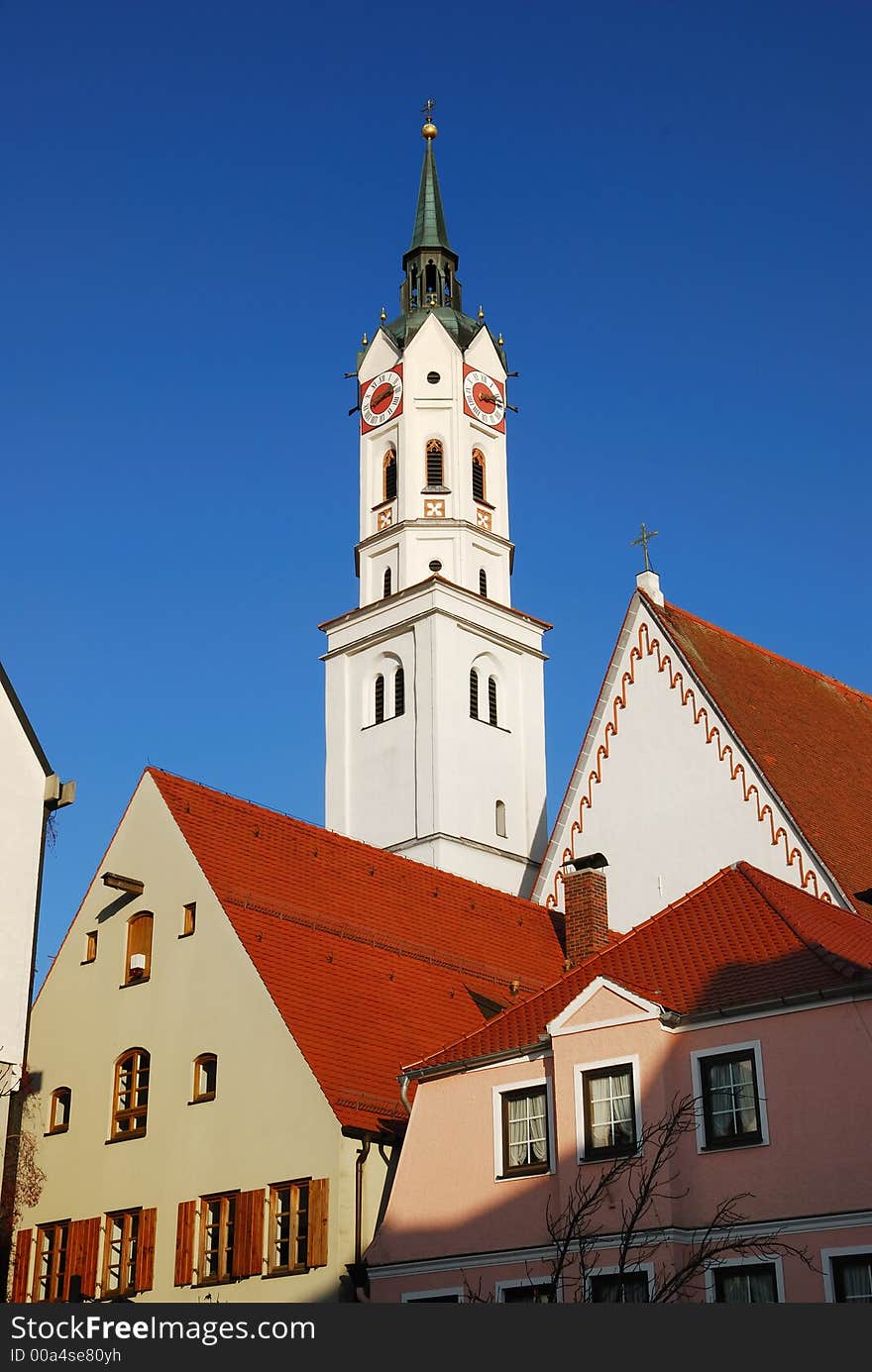 The image shows the white tower of an old gothic church in Bavaria. The top of the steeple is black, with a small ,golden cross on it. On the nave top you can see also a small cross. On each side of the tower is a clock, two of them can be seen in the picture. They are red with golden watch hands. It is a quarter past two. In foreground are some houses with red roofs. Some of the windows have shutters.  In the background of the image is deep blue sky.