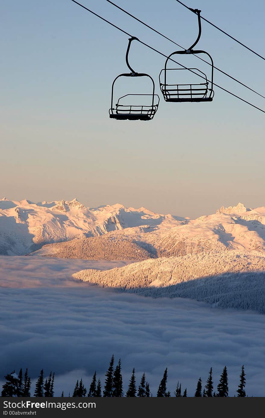 An empty chairlift takes riders high in the sky over mountains and valley clouds.