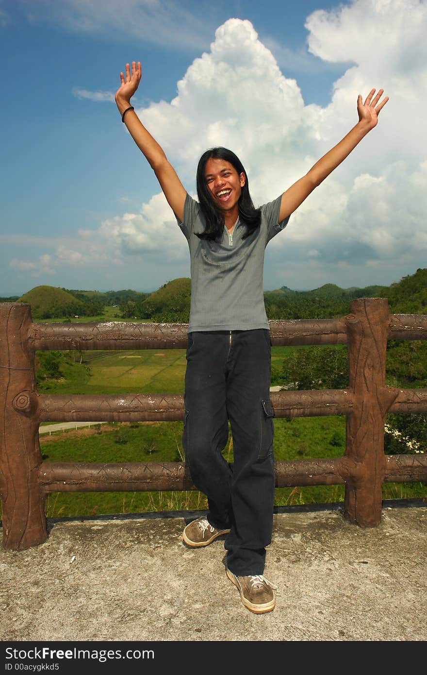 Asian exotic long-haired boy cheering, hands up, in front of the touristy landmark of the Chocolate Hills in Bohol, Philippines. Asian exotic long-haired boy cheering, hands up, in front of the touristy landmark of the Chocolate Hills in Bohol, Philippines.