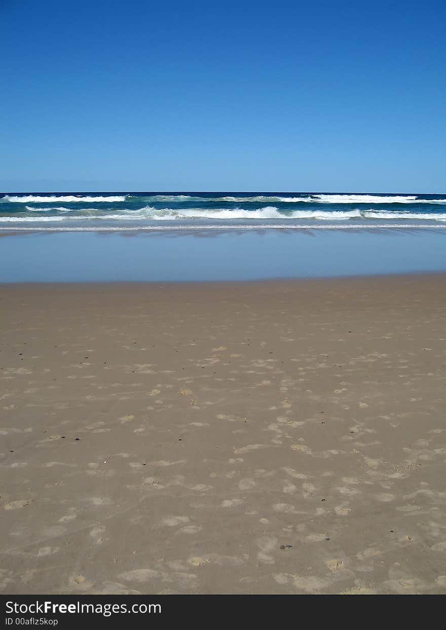 Beach at daytime. The sky is clear blue. Incoming waves from the sea. There are many foot prints on the sand.