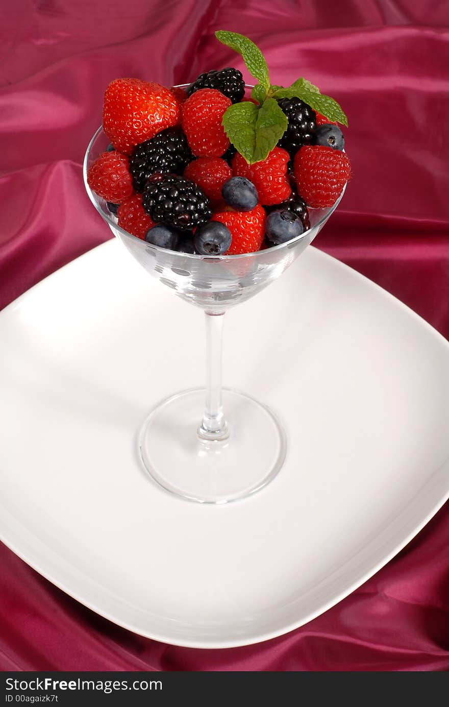 Fresh berries topped with mint in a martini glass resting on a white plate with a wine colored satin background