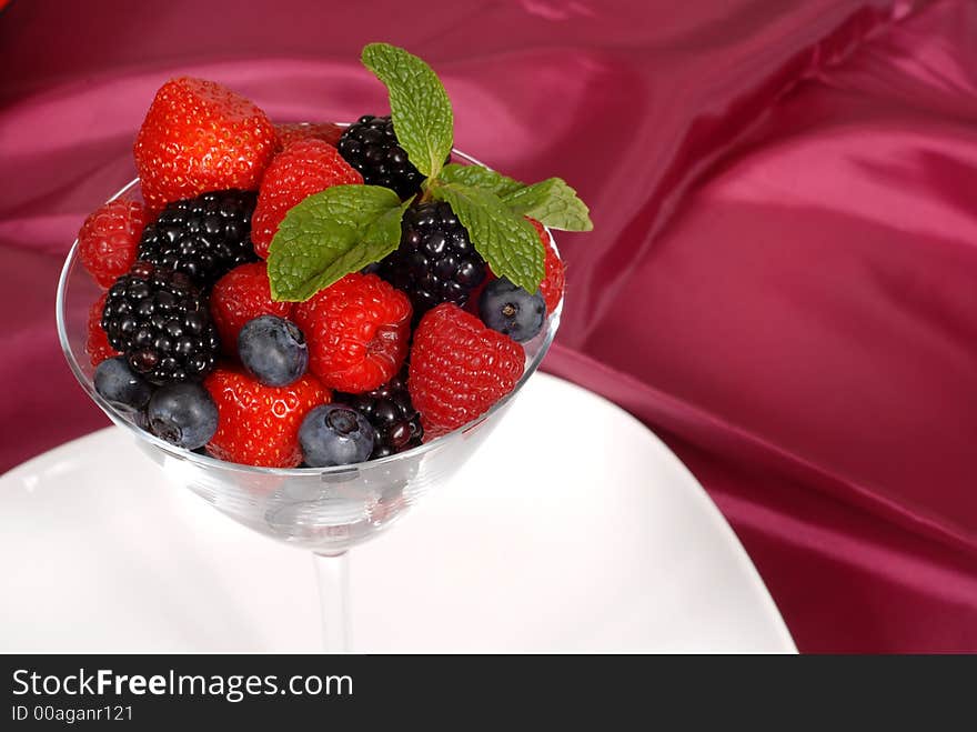 Close up of Fresh berries topped with mint in a martini glass resting on a white plate with a wine colored satin background