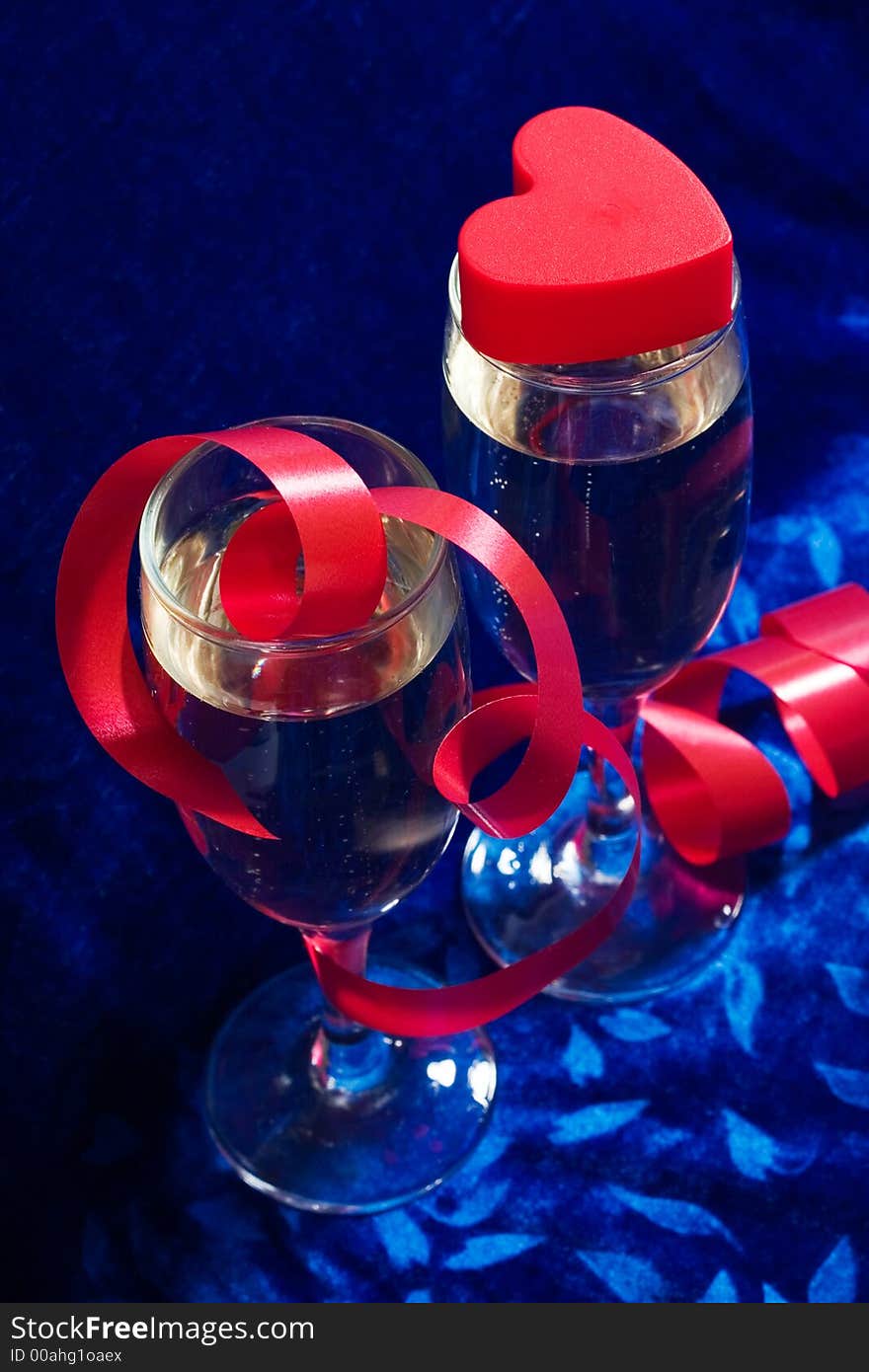 Two glasses of champagne, red twisted ribbon and plastic heart on the blue velvet. Two glasses of champagne, red twisted ribbon and plastic heart on the blue velvet