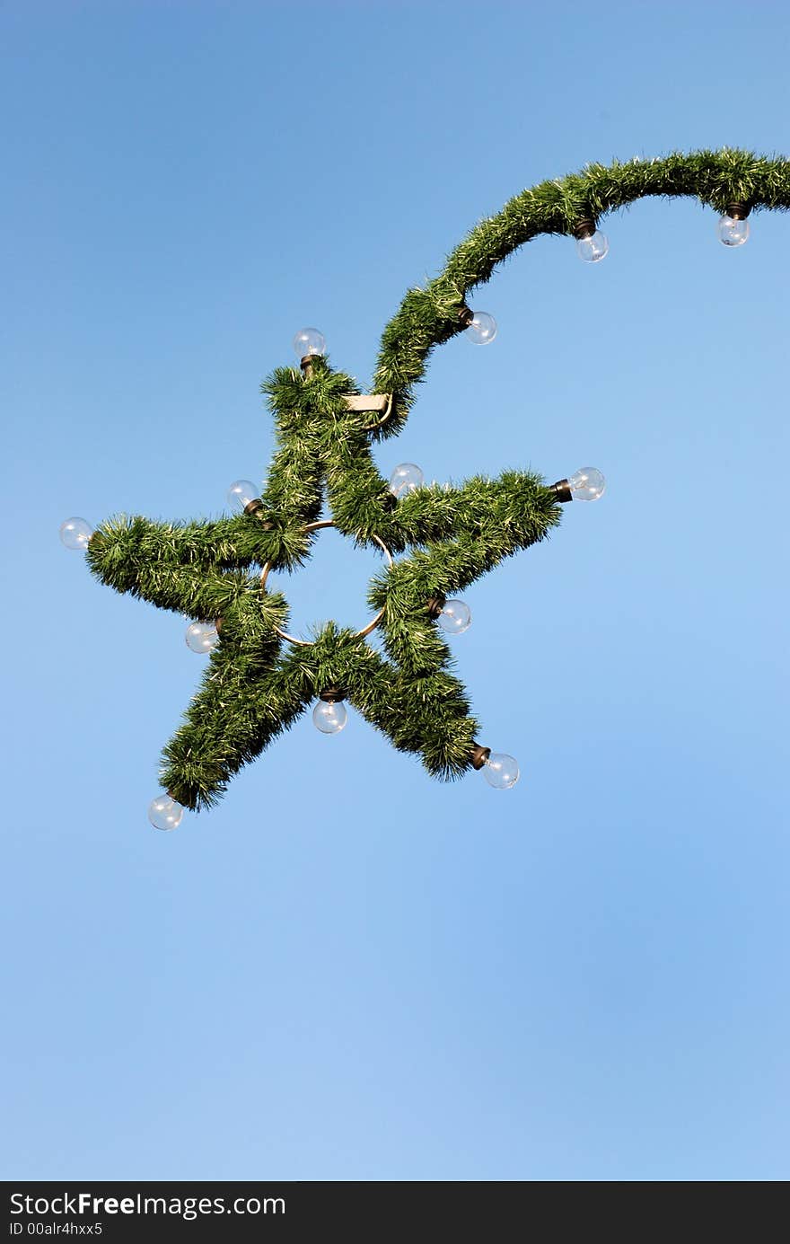 The picture shows a christmas star made of green fir branches. Attached are light bulbs.  In the background is blue sky. Image with copy space.