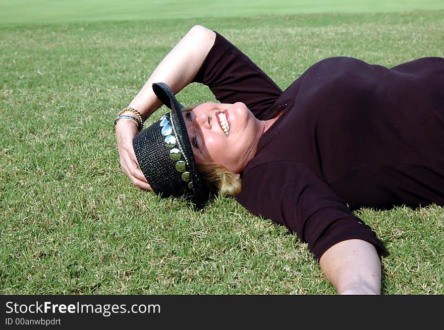 A smiling mature woman wearing a cowboy hat and lying down in the grass. Dressed in elbow length maroon sweater. Her left hand is holding her cowboy hat. . A smiling mature woman wearing a cowboy hat and lying down in the grass. Dressed in elbow length maroon sweater. Her left hand is holding her cowboy hat.