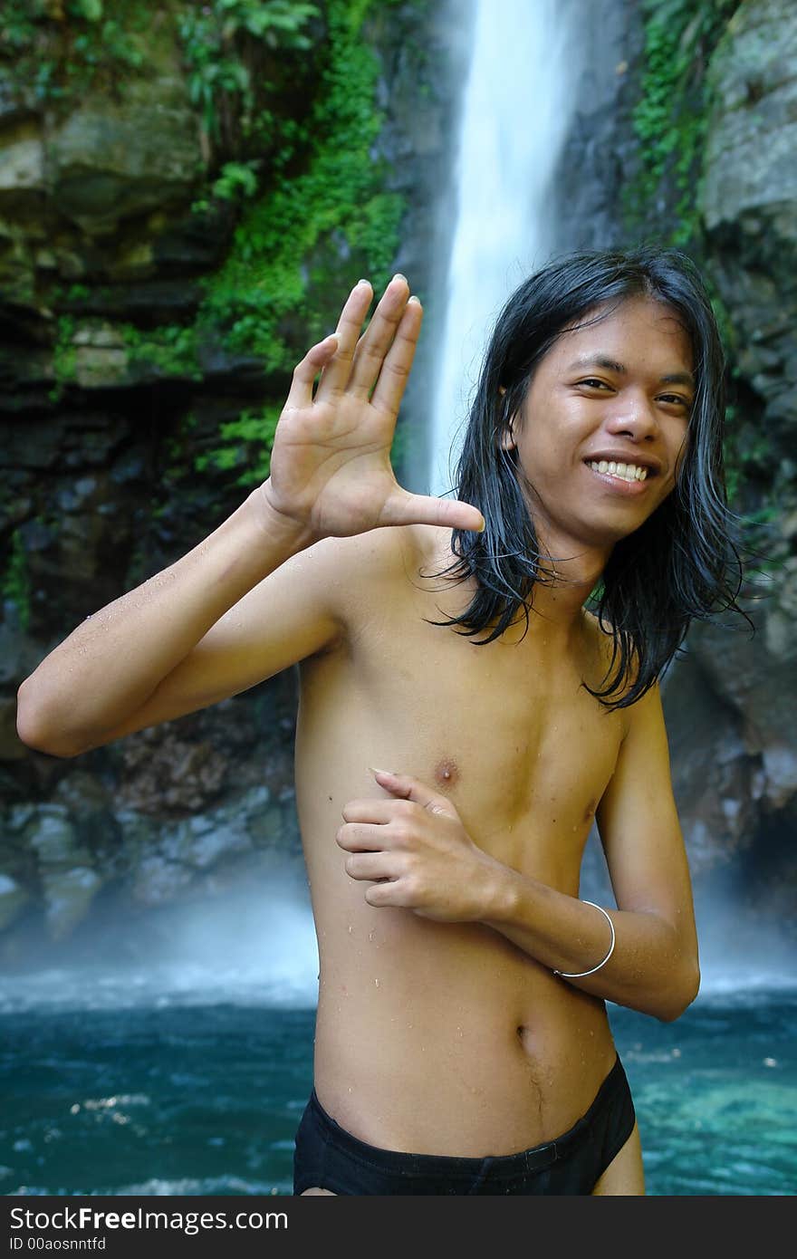 Asian exotic long-haired boy in front of a tropical pristine waterfall, after taking a dip in the whirlpool, waving and smiling. Concept of travel, tourism and outdoors. Asian exotic long-haired boy in front of a tropical pristine waterfall, after taking a dip in the whirlpool, waving and smiling. Concept of travel, tourism and outdoors.