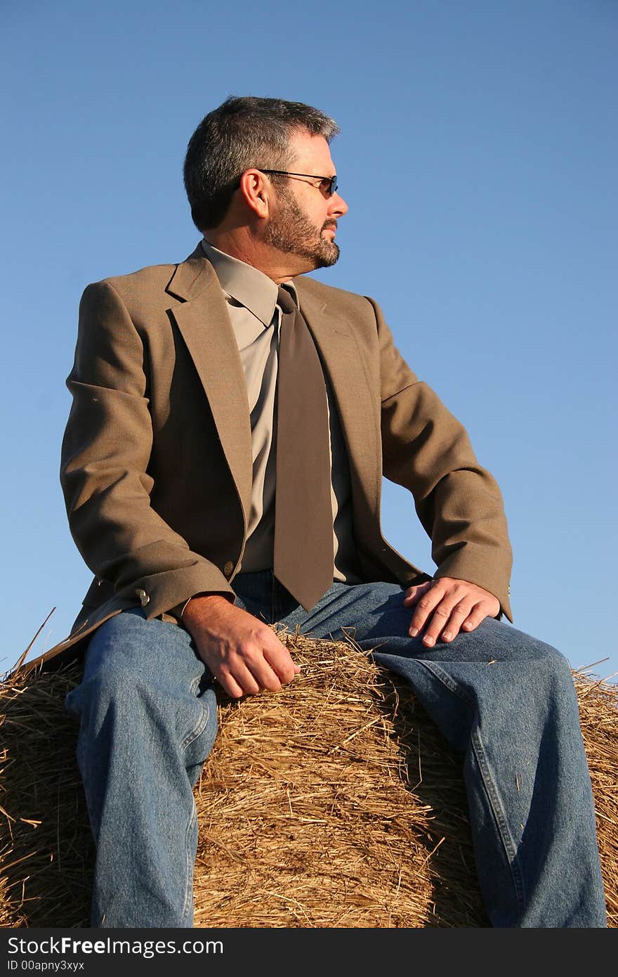 Man in jacket, tie and jeans sitting on a haystack.