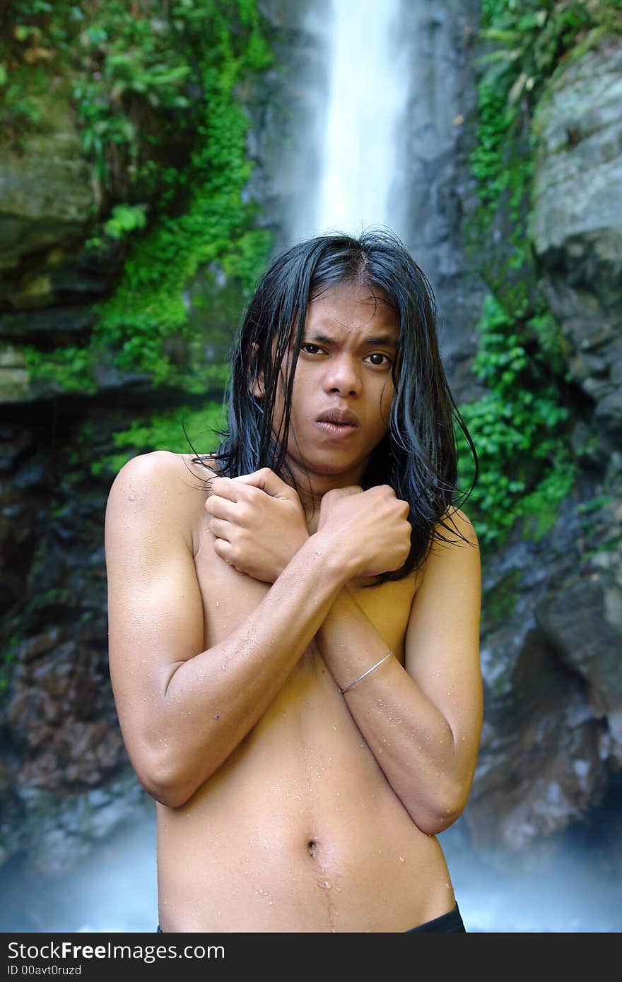 Asian exotic long-haired boy in front of a tropical pristine waterfall, after taking a dip in the whirlpool, shivering and covered with droplets. Asian exotic long-haired boy in front of a tropical pristine waterfall, after taking a dip in the whirlpool, shivering and covered with droplets.