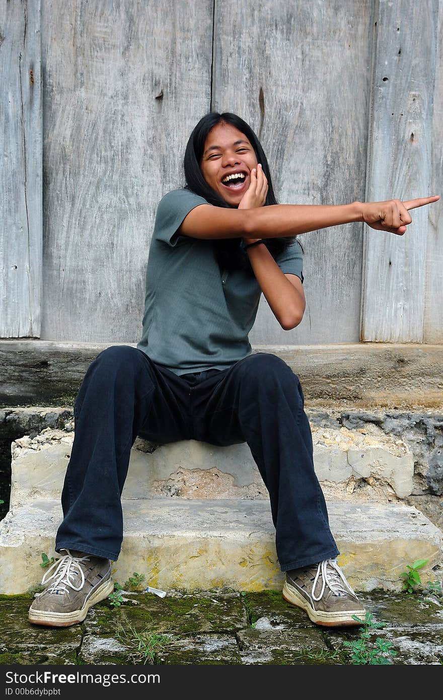 Asian tropical long-haired boy mockingly pointing with his finger in front of an old wooden weathered barn. Asian tropical long-haired boy mockingly pointing with his finger in front of an old wooden weathered barn.