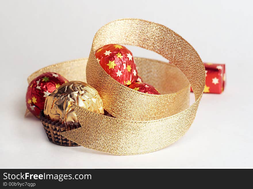 Chocolate golden sphere lay near a hill of sweets in a red metallic paper with golden stars on white background. Decorate of composition is golden strip. Chocolate golden sphere lay near a hill of sweets in a red metallic paper with golden stars on white background. Decorate of composition is golden strip.