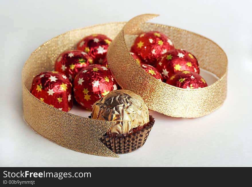 Chocolate golden sphere lay near a group of sweets in a red metallic paper with golden stars in the form of heard on white background. Decorate of composition is golden strip. Chocolate golden sphere lay near a group of sweets in a red metallic paper with golden stars in the form of heard on white background. Decorate of composition is golden strip.