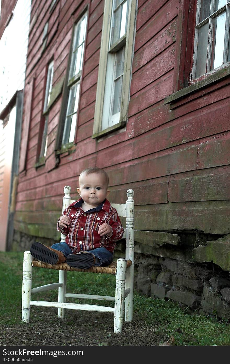 Image of baby boy sitting on a chair in front of a red farm building. Image of baby boy sitting on a chair in front of a red farm building