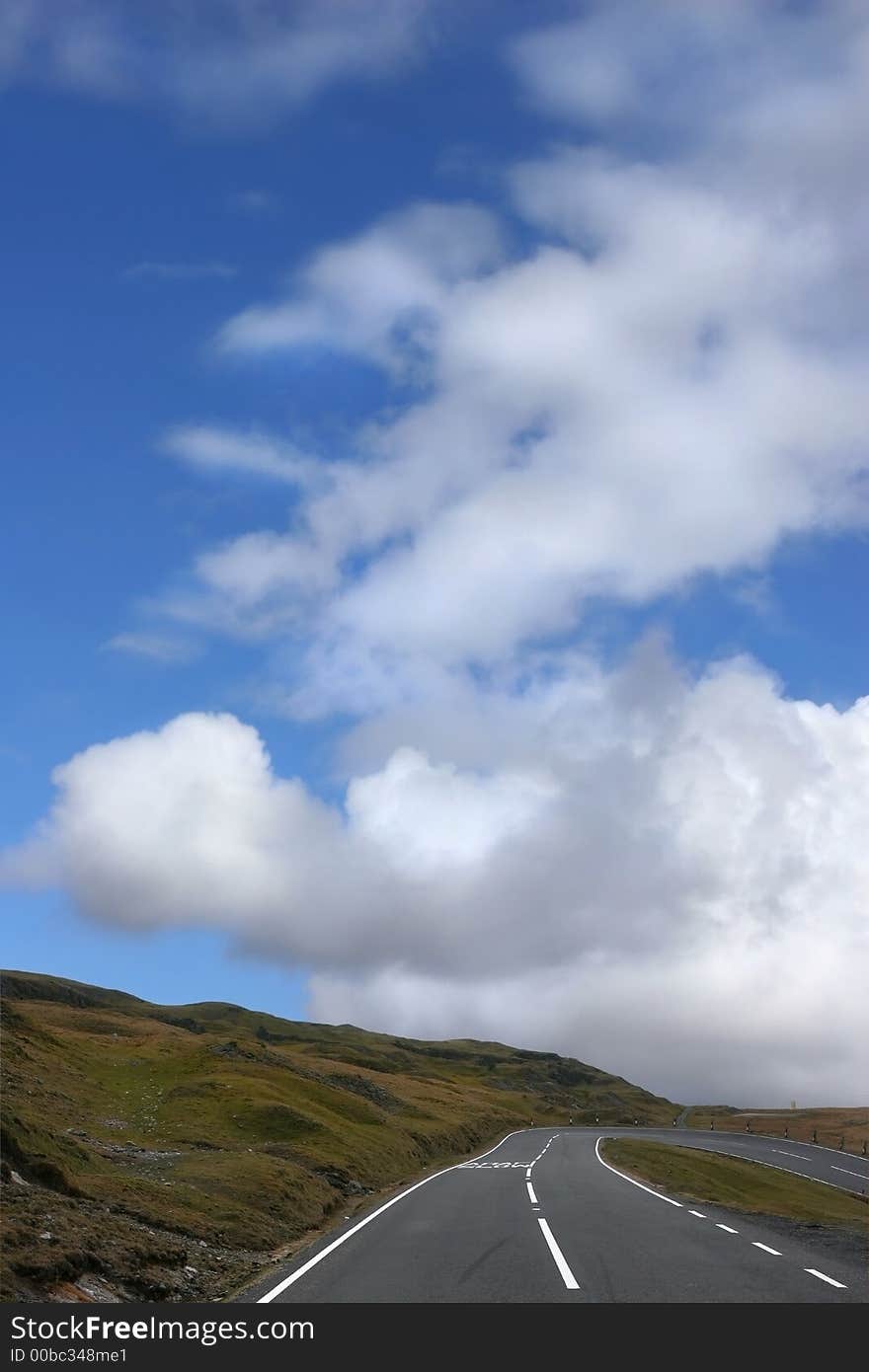 Empty down hill road in mountains with a dangerous right hand bend, with grass verges to either side, on a blue sky day with cumulus clouds. Located in the Brecon Beacons National Park, Wales, United Kingdom. Empty down hill road in mountains with a dangerous right hand bend, with grass verges to either side, on a blue sky day with cumulus clouds. Located in the Brecon Beacons National Park, Wales, United Kingdom.