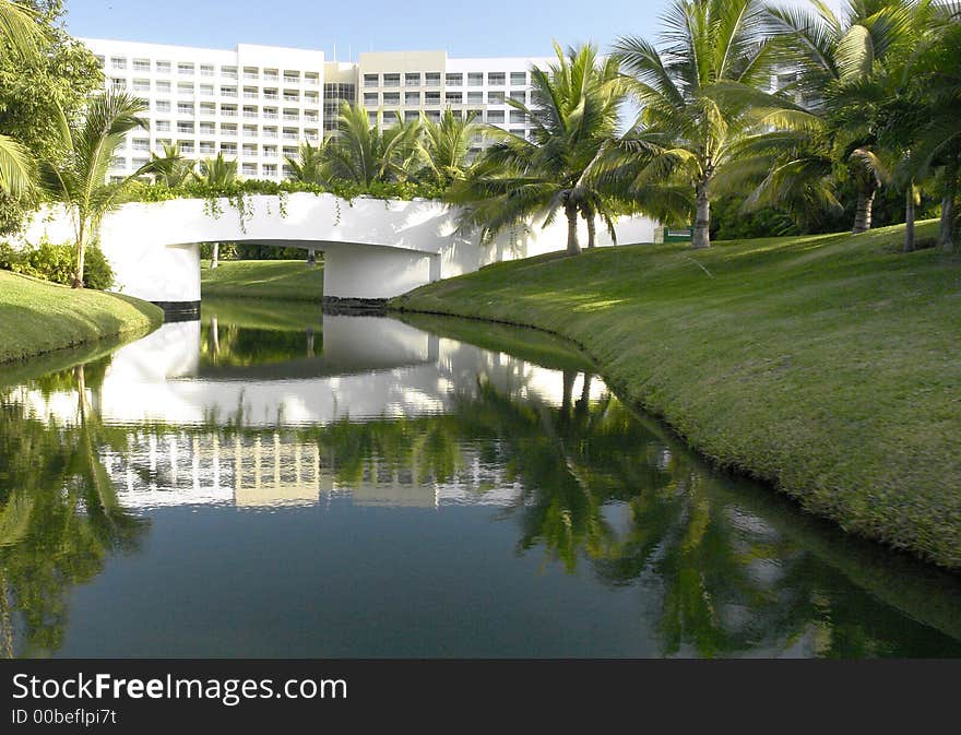 A reflection of a tropical resort with a water canal with a bridge in the forground and palm trees lining the canal with the resort in the background and  reflections of the resort in still waters