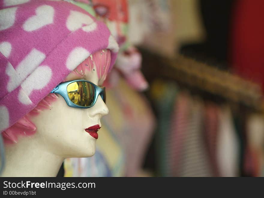 Old, worn mannequin with pink and white cap, sunglasses and deep red lipstick. Blurry, out-of-focus shop-background in the right two thirds of the photo. Old, worn mannequin with pink and white cap, sunglasses and deep red lipstick. Blurry, out-of-focus shop-background in the right two thirds of the photo.