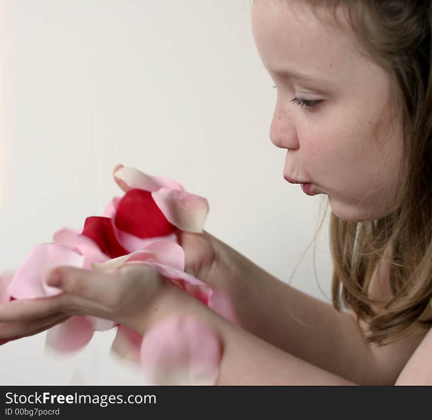 Young girl blowing pink, red, white rose petals held in hands over a slightly pink background. Young girl blowing pink, red, white rose petals held in hands over a slightly pink background