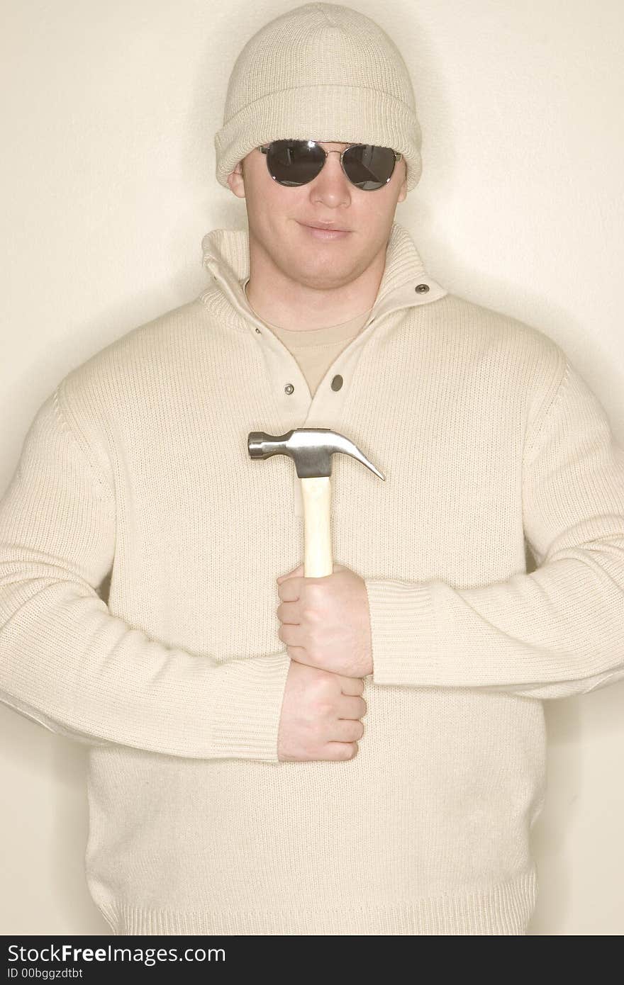 Man smiles as he holds a hammer to his chest as he wears sunglasses, a wool hat, and a wool sweater. Man smiles as he holds a hammer to his chest as he wears sunglasses, a wool hat, and a wool sweater