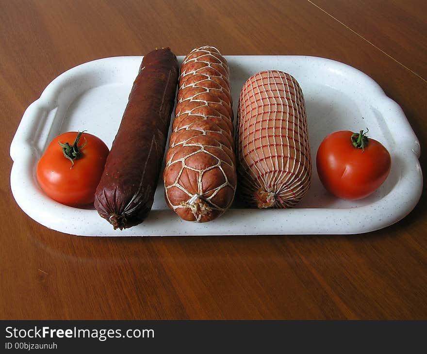 On a photo on a white tray 3 kinds of sausages, and from both sides two red tomatoes. The photo is made in Ukraine. On a photo on a white tray 3 kinds of sausages, and from both sides two red tomatoes. The photo is made in Ukraine.