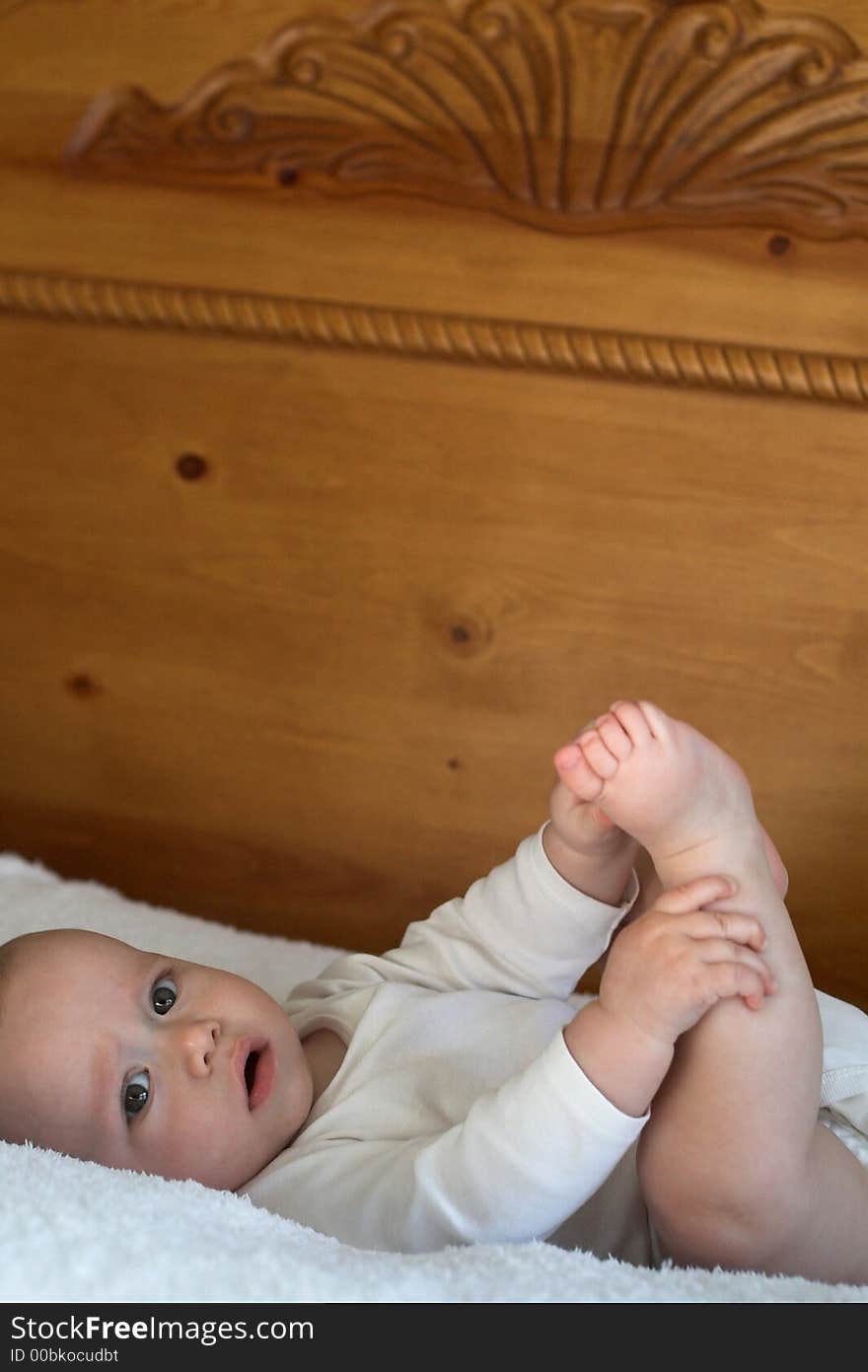 Image of baby boy wearing a white onesie lying on a bed playing with his feet. Image of baby boy wearing a white onesie lying on a bed playing with his feet