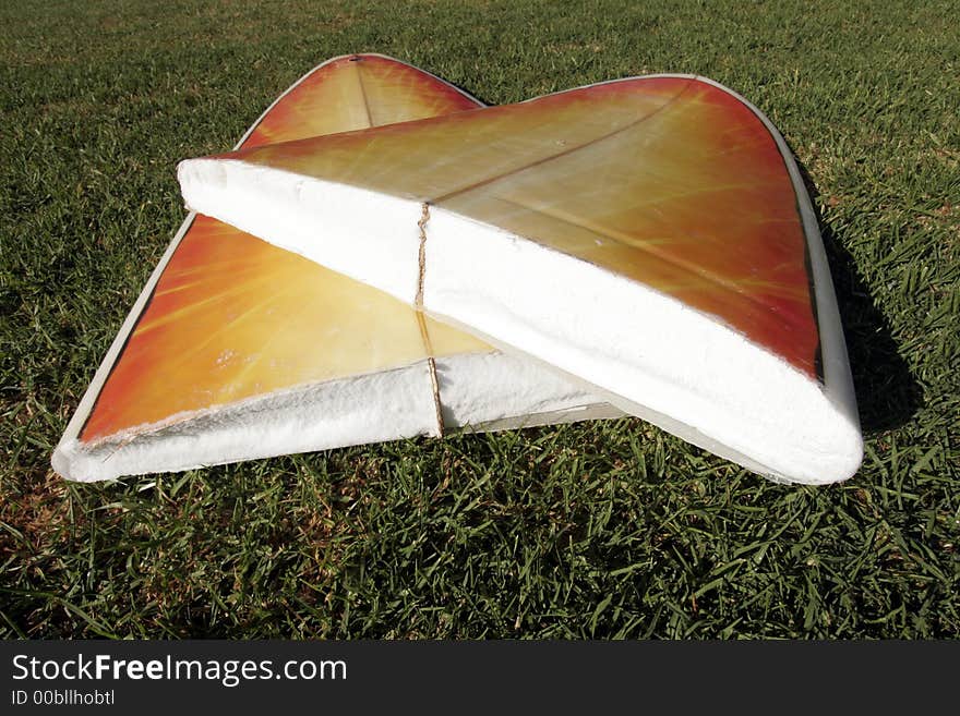 A Broken Yellow, Orange Surfboard On A Green Grass Field, Result Of Dangerous Surfing Conditions. A Broken Yellow, Orange Surfboard On A Green Grass Field, Result Of Dangerous Surfing Conditions