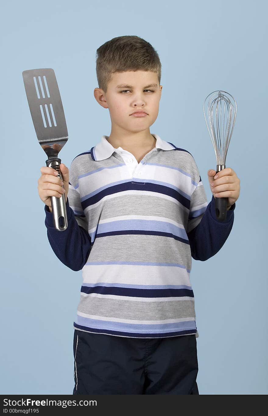 Young boy holding kitchen utensils with an angry expression on a blue background. Young boy holding kitchen utensils with an angry expression on a blue background.