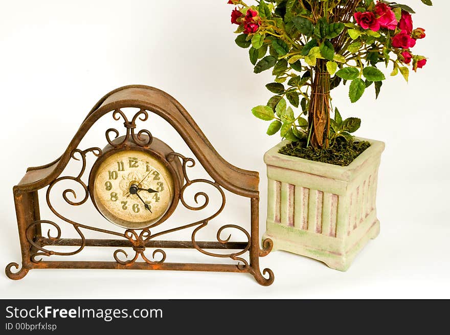 A rusty and dusty old wire-frame clock sits in front of a decorative pot of miniature red roses on a white background. A rusty and dusty old wire-frame clock sits in front of a decorative pot of miniature red roses on a white background.