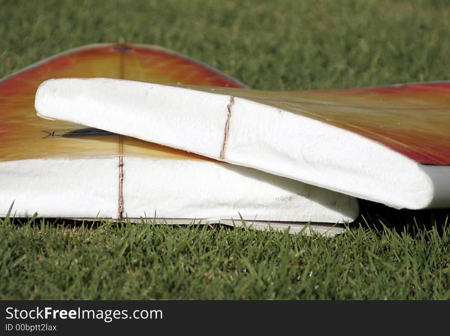 A Broken Yellow, Orange Surfboard On A Green Grass Field, Result Of Dangerous Surfing Conditions, Shallow Depth Of Field. A Broken Yellow, Orange Surfboard On A Green Grass Field, Result Of Dangerous Surfing Conditions, Shallow Depth Of Field