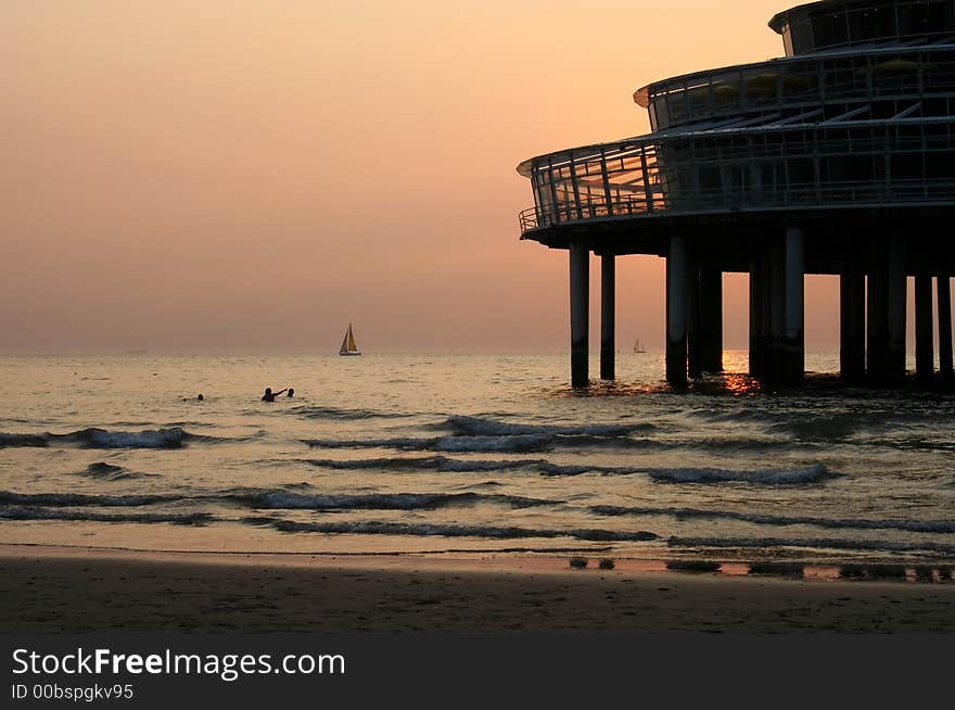 Sea with swimmers in the sunset, with a pier restaurant and a sailing boat on the horizon. Sea with swimmers in the sunset, with a pier restaurant and a sailing boat on the horizon