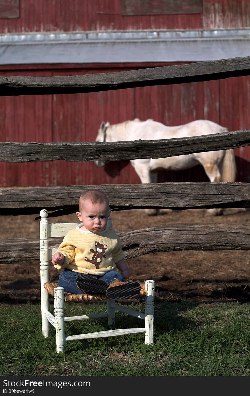 Image of baby boy sitting on a chair in front of a barn building, a horse in the background. Image of baby boy sitting on a chair in front of a barn building, a horse in the background