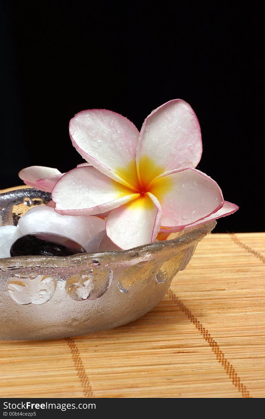 Detail of frangipane flower and pebbles in a glass bowl on the rattan background. Detail of frangipane flower and pebbles in a glass bowl on the rattan background