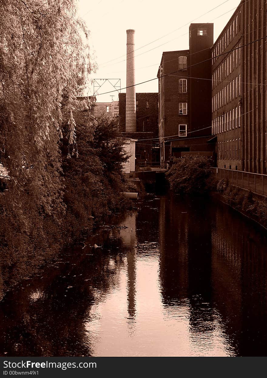 Sepia image of canal behind old factory building in easthampton massachusetts, on the left is a bank of trees, on the right a bank of factory windows, in the center a smoke stack. Sepia image of canal behind old factory building in easthampton massachusetts, on the left is a bank of trees, on the right a bank of factory windows, in the center a smoke stack