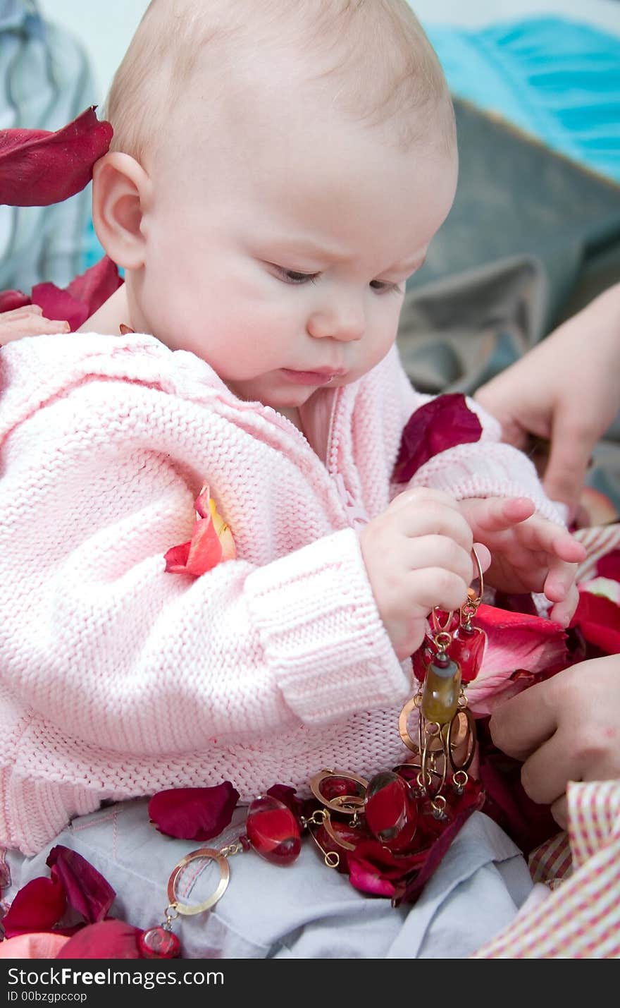 Cute baby girl sitting and looking to the red beads in her hands. Roses petals are around her. Cute baby girl sitting and looking to the red beads in her hands. Roses petals are around her.