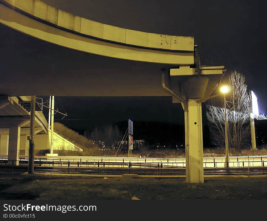 A traffic bridge over tracks at night, with one electric light in front of a tree.