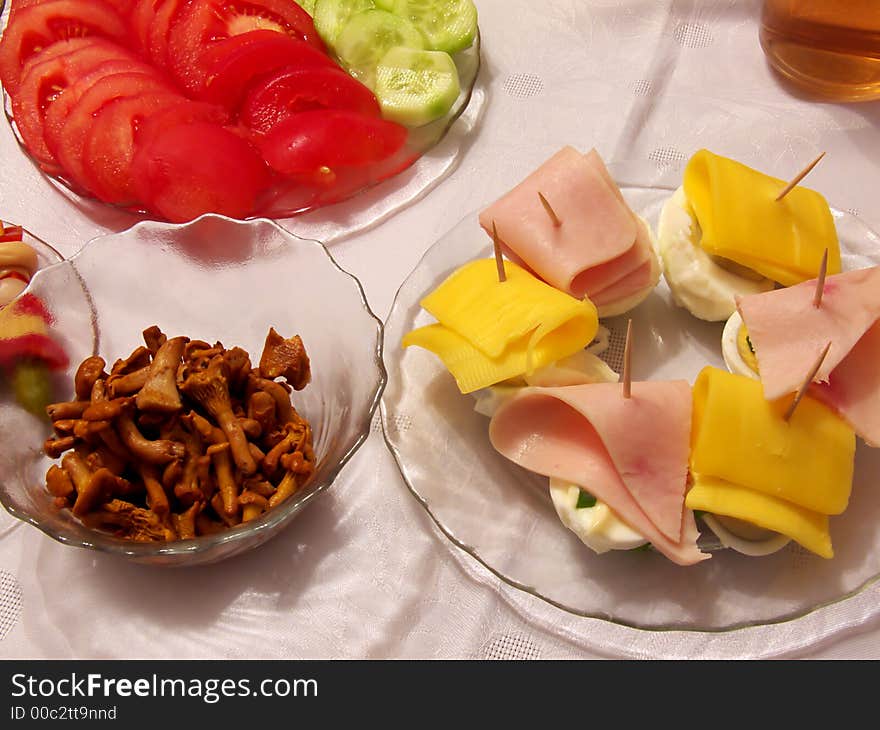 Piece of holiday supper table: tomato, cucumber, pickled cucumbers, cork, red paprika, onion, fungus, egg, cheese, ham, on the plates, all and cup of tea on the tablecloth