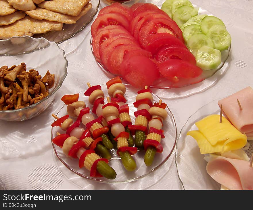 Piece of holiday supper table:
tomato, cucumber, pickled cucumbers, cork, red paprika, onion, fungus, egg, cheese, ham, poultry chop on the plates, 
all on the tablecloth